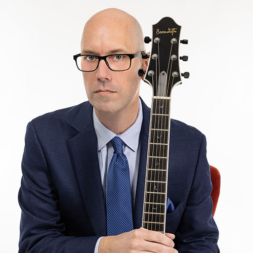 Shawn Purcell in a navy suit holding the neck of his guitar
