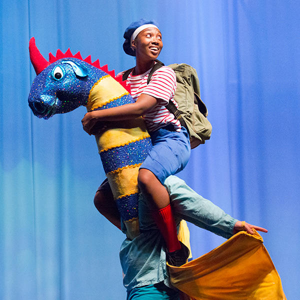 A young African American wearing a blue hair cap, red and white stripe shirt, blue shorts, red tall socks, and army green backpack riding a puppet dinosaur.